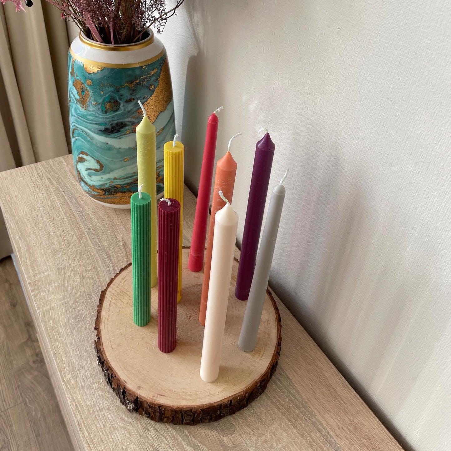 Colorful taper candles for dinner table displayed on wooden slice with decorative vase background